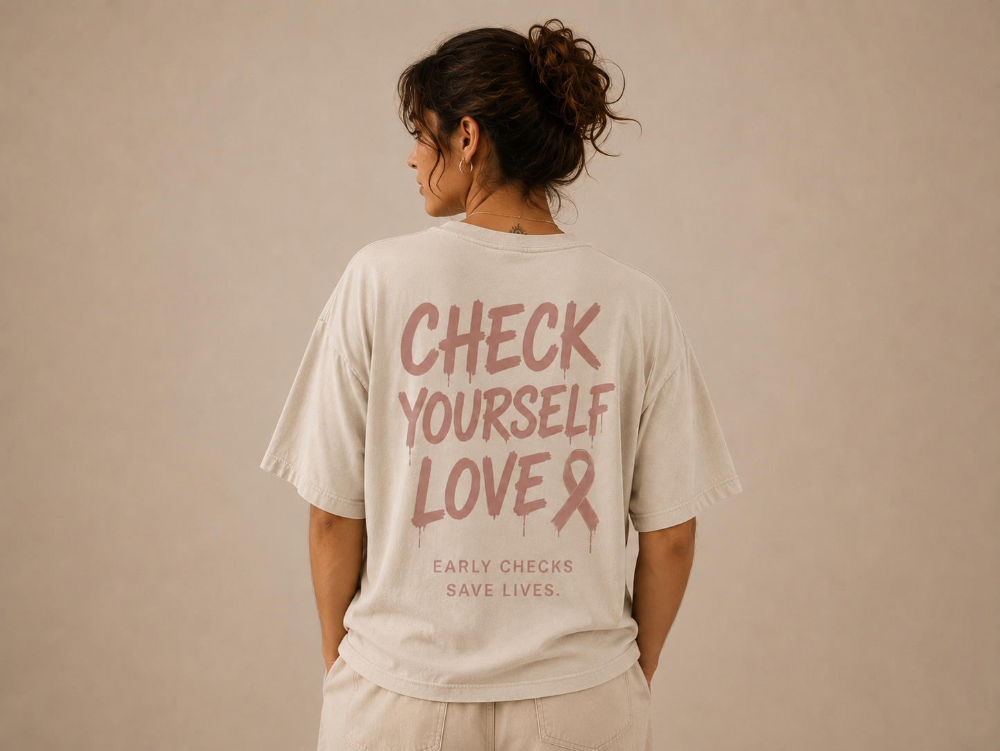 Rückenansicht eines cremefarbenen Oversize-Shirts mit dem Print „Check yourself love — Early checks save lives“ zum Thema Brustkrebsvorsorge